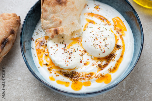 Bowl of turkish poached eggs served in yogurt and topped with melted spicy butter, middle closeup on a beige stone background