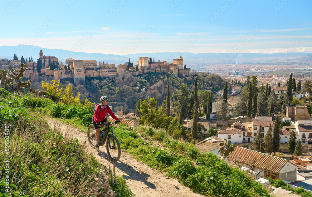 nice active senior woman cycling with her electric mountain bike in Granada below the world heritage site of Alhambra, Granada, Andalusia,  Spain - obrazy, fototapety, plakaty 