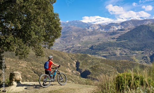 nice, active senior woman cycling with her electric mountain bike below the snow covered mountains of the Spanish Sierra Nevada, near Granada, Andalusia, Spain