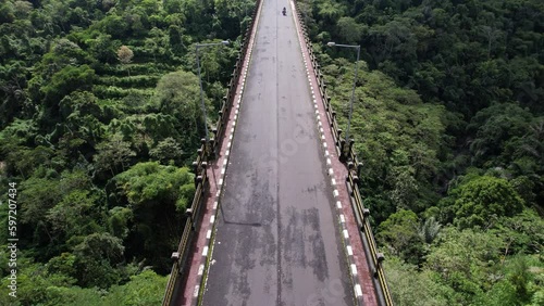 Lengthwise view of elevated roadway, aerial shot of Tukad Bangkung bridge. Camera fly along road and tilt up, green tropical plants seen at slope of large ravine. One motorbike ride by at empty way photo