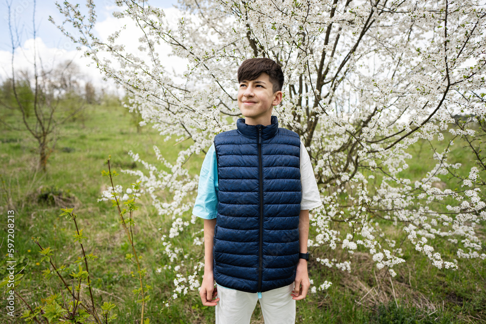 Fototapeta premium Teen boy wear sleeveless vest against white blloming tree in spring.