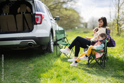 Mother with two daughters sit on chair against car open trunk on picnic.