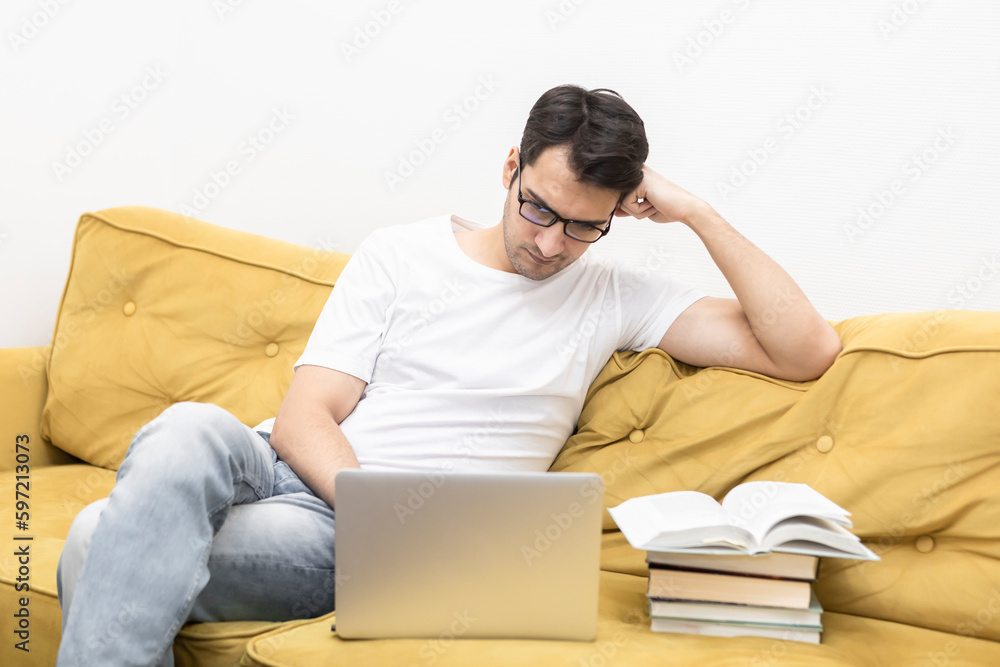 Confused man sitting on the couch with a lot of books. Online learning and home education concept