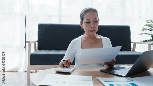 Confident asian chinese mature businesswoman working with laptop and desktop computer doing some paperwork at office workplace.