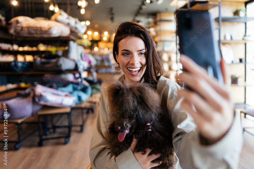Beautiful and happy young woman sitting in modern pet shop cafe bar and enjoying in fresh coffee together with her adorable brown toy poodle. She also taking self portrait photo with smart phone.