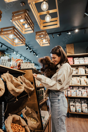 Beautiful young woman enjoying in modern pet shop together with her adorable brown toy poodle.