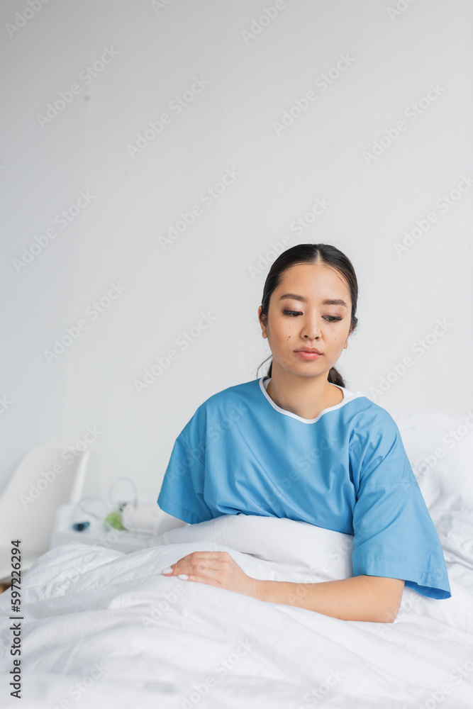 depressed asian woman in hospital gown thinking while sitting on bed in clinic. depressed asian woman in hospital gown thinking while sitting on bed in clinic.