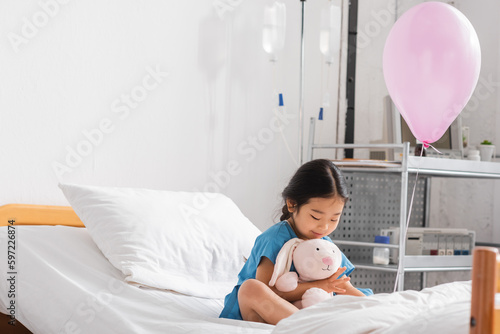cheerful asian girl playing with toy bunny near festive balloon on hospital bed.