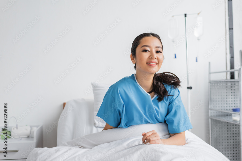 cheerful asian woman sitting on bed in hospital ward and smiling at camera. cheerful asian woman sitting on bed in hospital ward and smiling at camera.