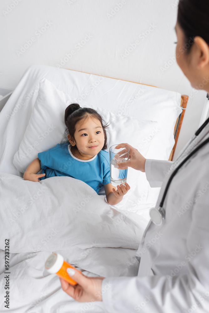 cheerful asian child taking glass of water from doctor holding pills container in hospital ward. cheerful asian child taking glass of water from doctor holding pills container in hospital ward.