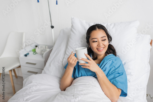 pleased asian woman lying on hospital bed with cup of tea and looking away.
