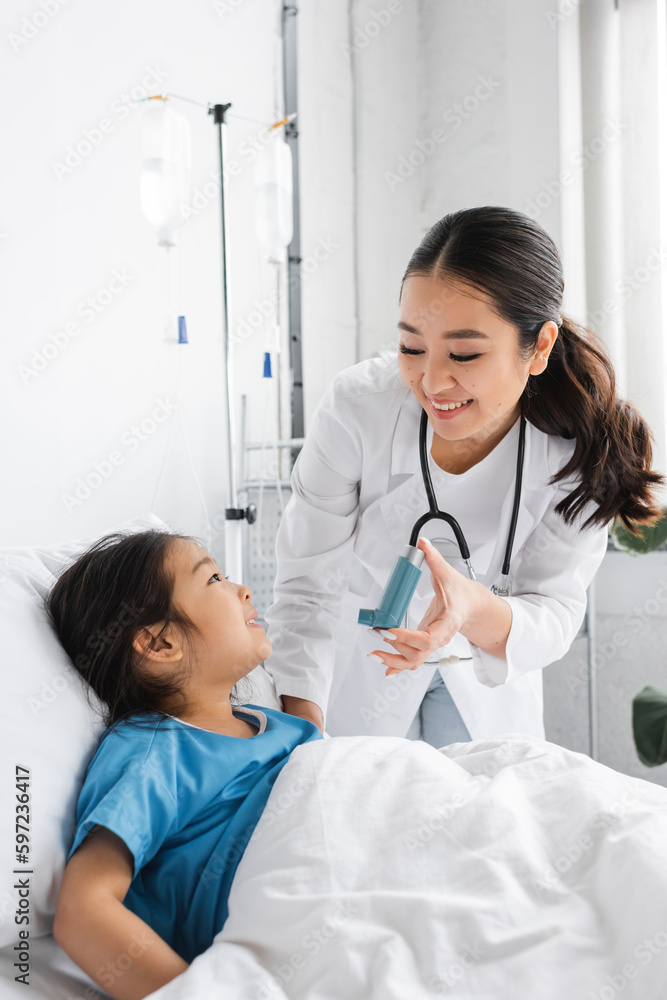 smiling doctor holding inhaler near positive girl on bed in hospital ward. smiling doctor holding inhaler near positive girl on bed in hospital ward.