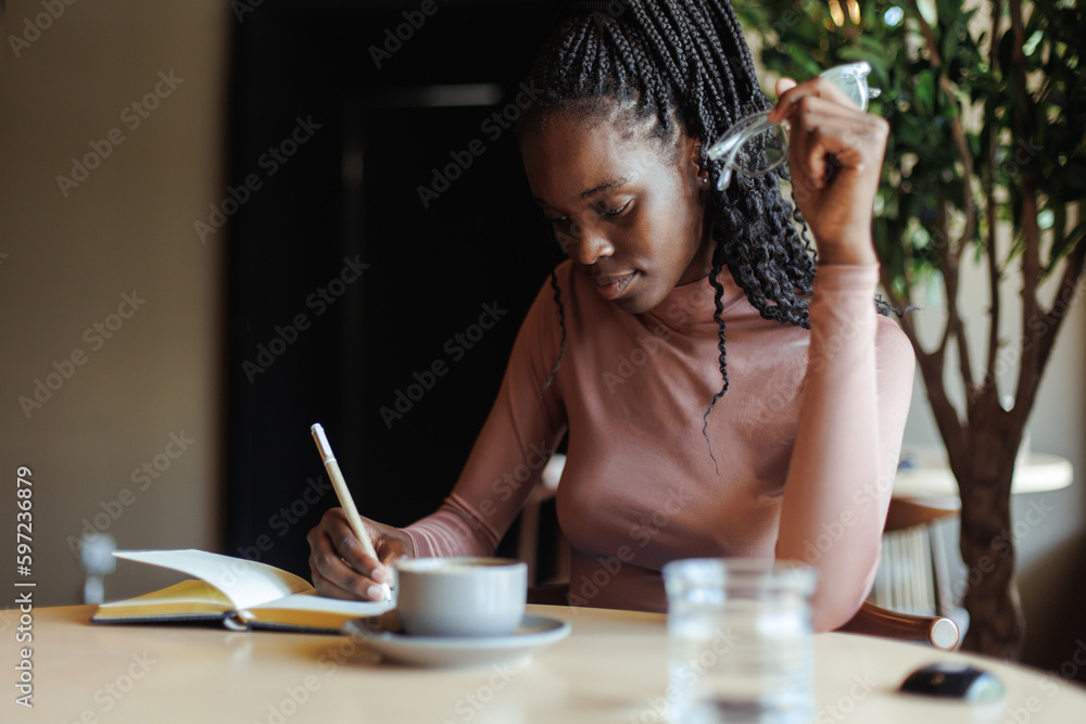 Portrait of young focused African-American woman with long dark braids wearing pink roll-neck sweater, sitting at beige table with cup of coffee in cafe, writing text in notebook. Education, diary. Portrait of young focused African-American woman with long dark braids wearing pink roll-neck sweater, sitting at beige table with cup of coffee in cafe, writing text in notebook. Education, diary.