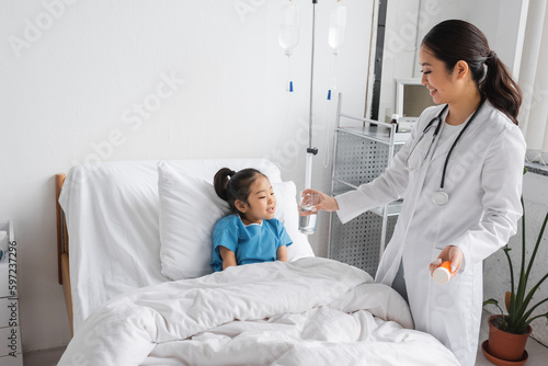 smiling pediatrician holding pills container and glass of water near carefree asian girl on bed in clinic.