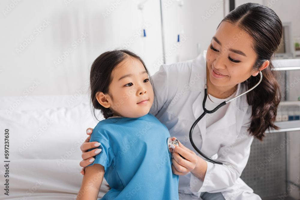joyful doctor examining little asian girl with stethoscope in pediatric clinic.