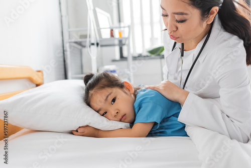 young asian doctor calming sad child lying on bed in clinic.
