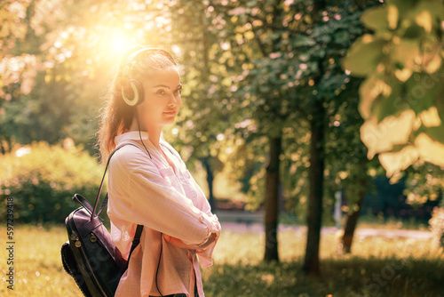 Lonely young woman wearing headset listening music in urban park at sunset. Female person walking with headphones outdoor.
