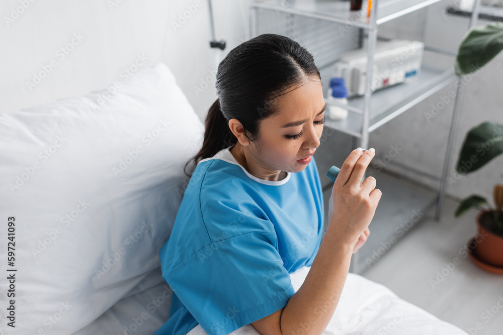 high angle view of sick asian woman holding inhaler while sitting on bed in hospital ward.