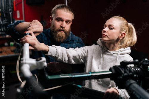 Professional bearded cycling repairman having conversation with blonde female client, talking about problem of bicycle detected during diagnostics in repair shop with dark interior.