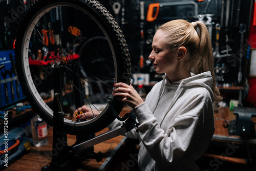Portrait of pretty blonde female cycling repairman checking bicycle wheel spoke with bike spoke key in repair workshop with dark interior. Concept of professional maintenance of bicycle transport.