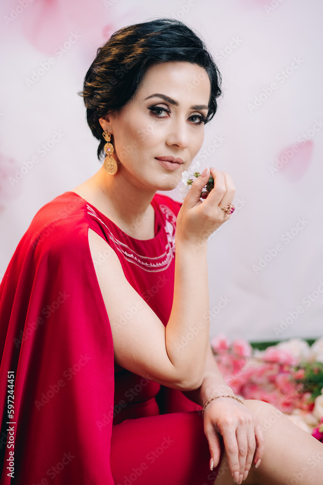 Luxurious woman in red dress sitting on the floor posing for camera and holding blooming flower