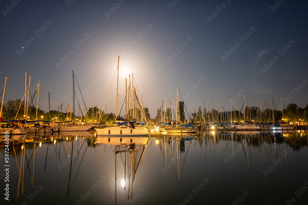 Night view of Balaton lake in Hungary, scenic landscape with yacht marine in moonlight, starry sky and reflection in the water, outdoor travel background