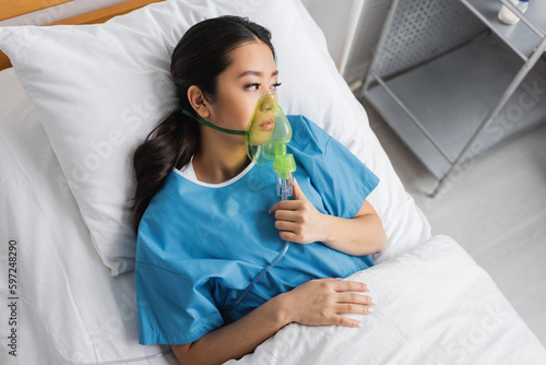 top view of upset asian woman lying on hospital bed in oxygen mask and looking away. photo