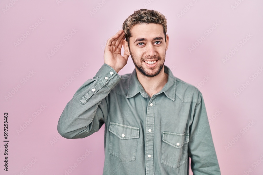 Hispanic man with beard standing over pink background smiling with hand over ear listening an hearing to rumor or gossip. deafness concept. Hispanic man with beard standing over pink background smiling with hand over ear listening an hearing to rumor or gossip. deafness concept.