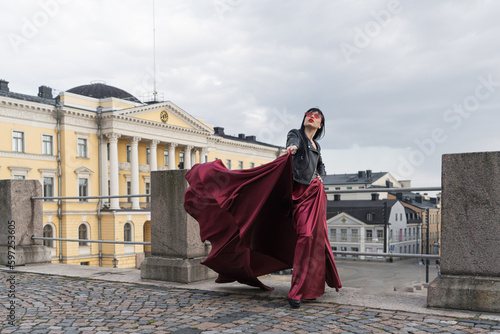 Helsinki. Finland. 25.04.2023 .A sexy woman with a perfect figure stands on Helsinki's Central Square, wearing a red satin skirt and a leather biker jacket. Her skirt billows in the wind