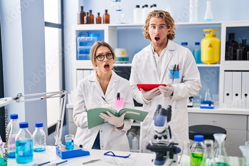 Two people working at scientist laboratory in shock face  looking skeptical and sarcastic  surprised with open mouth
