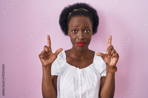 African woman with curly hair standing over pink background pointing up looking sad and upset, indicating direction with fingers, unhappy and depressed.