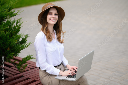 Summer portrait of a female freelancer in light clothes and a hat sitting on a bench with a laptop in a summer park outdoors, working on a laptop.The concept of urban lifestyle, technology.Copy space.