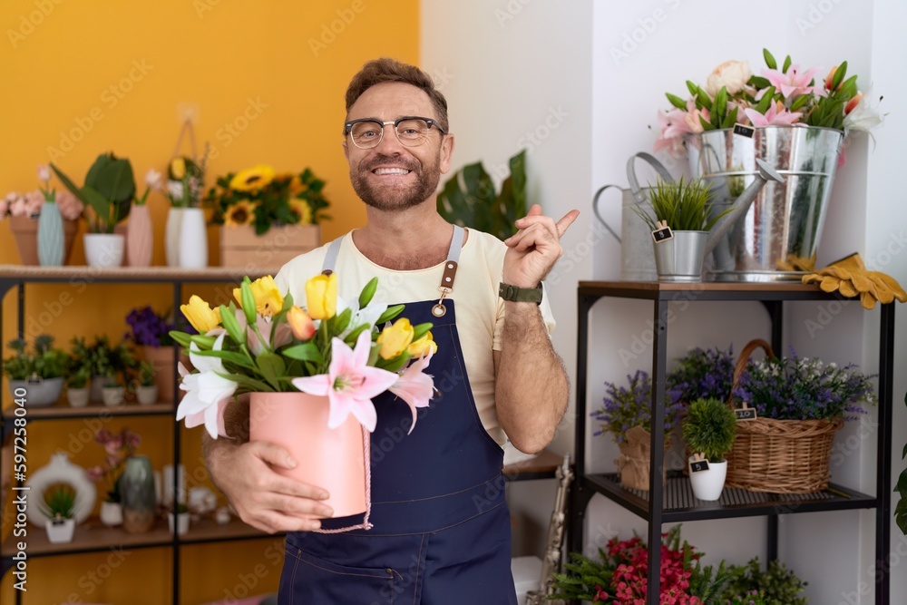 Middle age man with beard working at florist shop holding plant smiling happy pointing with hand and finger to the side Middle age man with beard working at florist shop holding plant smiling happy pointing with hand and finger to the side