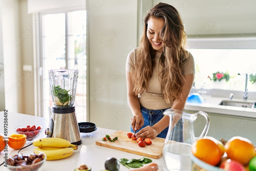 Young beautiful hispanic woman preparing vegetable smoothie with blender cutting strawberries at the kitchen Young beautiful hispanic woman preparing vegetable smoothie with blender cutting strawberries at the kitchen