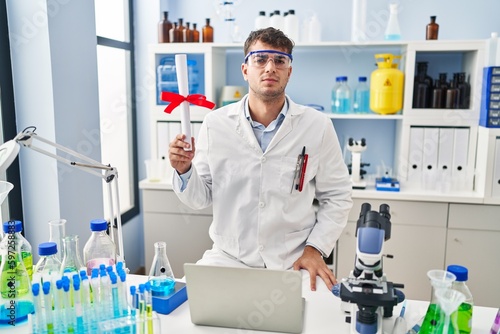 Young hispanic man working at scientist laboratory holding diploma thinking attitude and sober expression looking self confident