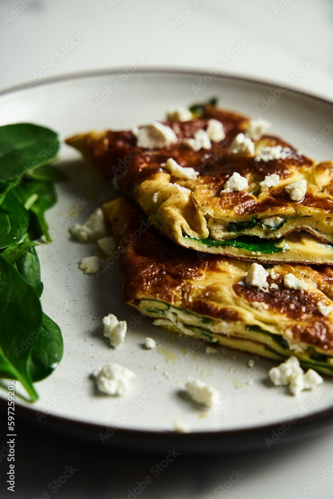 Close up of sliced folded omelette stuffed with spinach and feta on the white plate with spinach leaves on back. Healthy and dietary breakfast dish. Minimalistic food. White marble background. 