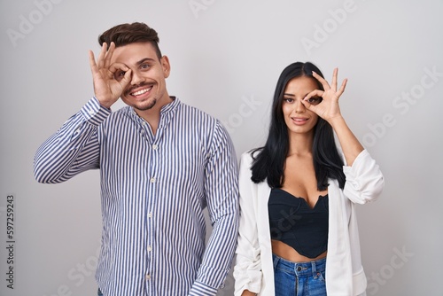 Young hispanic couple standing over white background doing ok gesture with hand smiling, eye looking through fingers with happy face.