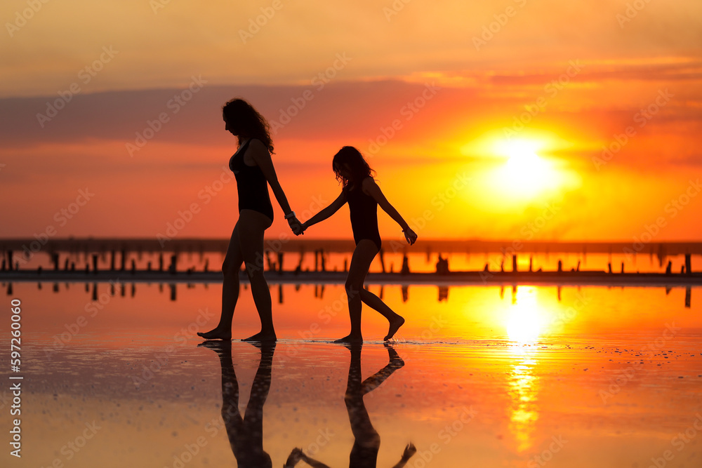 Happy family summer travel holiday. Silhouette of mom with child daughter holding hands walking together on beach on sunset. Happy mothers day. Concept of family values. International Children's Day