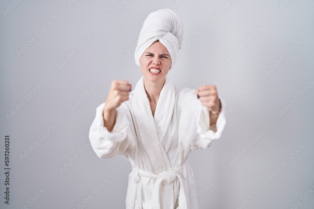 Blonde caucasian woman wearing bathrobe angry and mad raising fists frustrated and furious while shouting with anger. rage and aggressive concept. Blonde caucasian woman wearing bathrobe angry and mad raising fists frustrated and furious while shouting with anger. rage and aggressive concept.