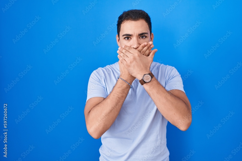 Young hispanic man standing over blue background shocked covering mouth with hands for mistake. secret concept. Young hispanic man standing over blue background shocked covering mouth with hands for mistake. secret concept.