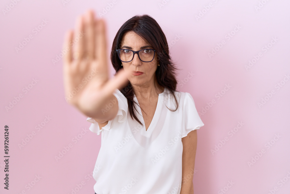 Middle age hispanic woman wearing casual white t shirt and glasses doing stop sing with palm of the hand. warning expression with negative and serious gesture on the face. Middle age hispanic woman wearing casual white t shirt and glasses doing stop sing with palm of the hand. warning expression with negative and serious gesture on the face.