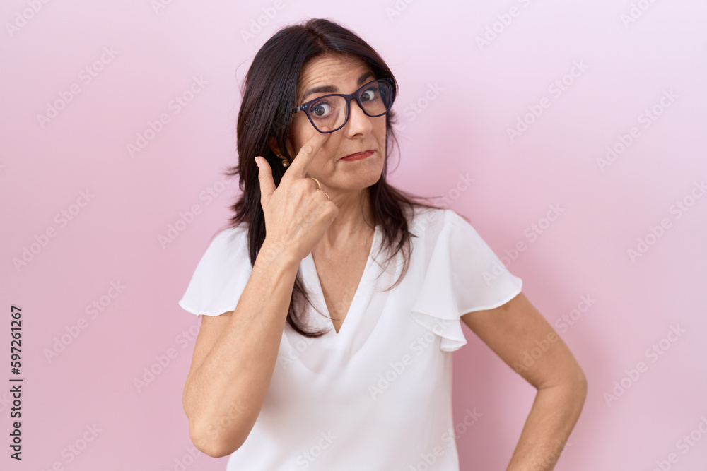 Middle age hispanic woman wearing casual white t shirt and glasses pointing to the eye watching you gesture, suspicious expression Middle age hispanic woman wearing casual white t shirt and glasses pointing to the eye watching you gesture, suspicious expression