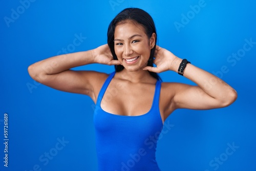 Hispanic woman standing over blue background relaxing and stretching, arms and hands behind head and neck smiling happy © Krakenimages.com
