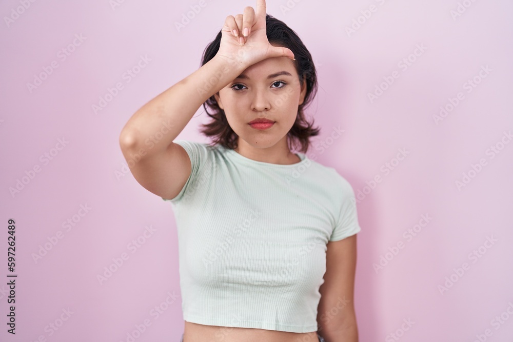 Hispanic young woman standing over pink background making fun of people with fingers on forehead doing loser gesture mocking and insulting. Hispanic young woman standing over pink background making fun of people with fingers on forehead doing loser gesture mocking and insulting.