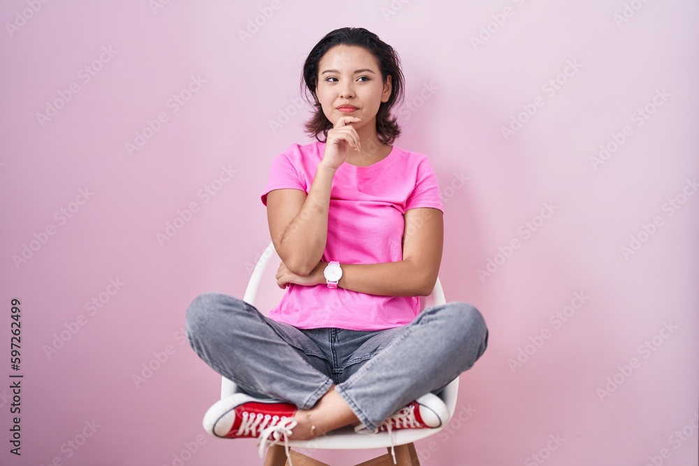 Hispanic young woman sitting on chair over pink background with hand on chin thinking about question, pensive expression. smiling and thoughtful face. doubt concept. Hispanic young woman sitting on chair over pink background with hand on chin thinking about question, pensive expression. smiling and thoughtful face. doubt concept.