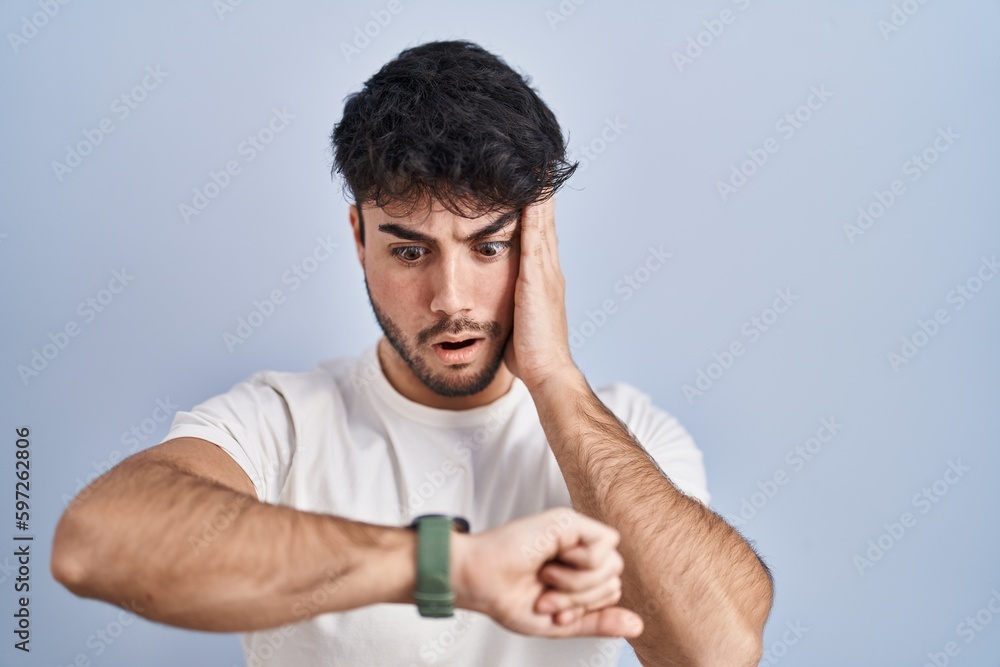 Hispanic man with beard standing over white background looking at the watch time worried, afraid of getting late Hispanic man with beard standing over white background looking at the watch time worried, afraid of getting late