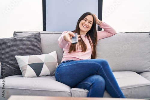 Young brunette woman holding television remote control looking positive and happy standing and smiling with a confident smile showing teeth