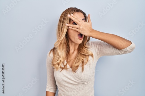 Young blonde woman standing over isolated background peeking in shock covering face and eyes with hand, looking through fingers with embarrassed expression.