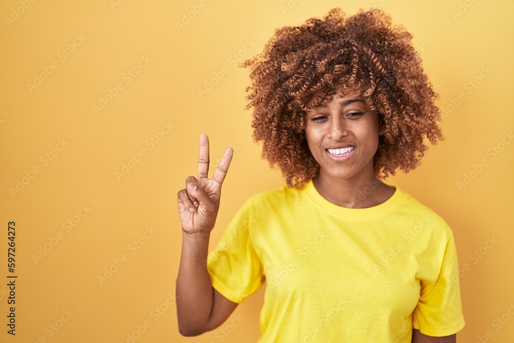 Young hispanic woman with curly hair standing over yellow background showing and pointing up with fingers number two while smiling confident and happy. Young hispanic woman with curly hair standing over yellow background showing and pointing up with fingers number two while smiling confident and happy.