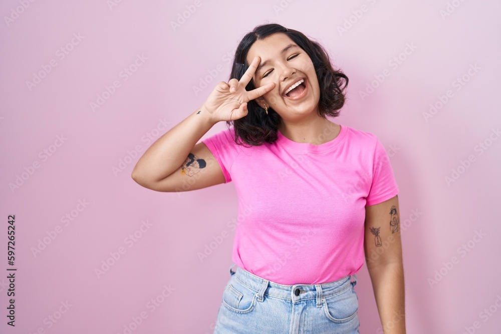 Young hispanic woman standing over pink background doing peace symbol with fingers over face, smiling cheerful showing victory Young hispanic woman standing over pink background doing peace symbol with fingers over face, smiling cheerful showing victory